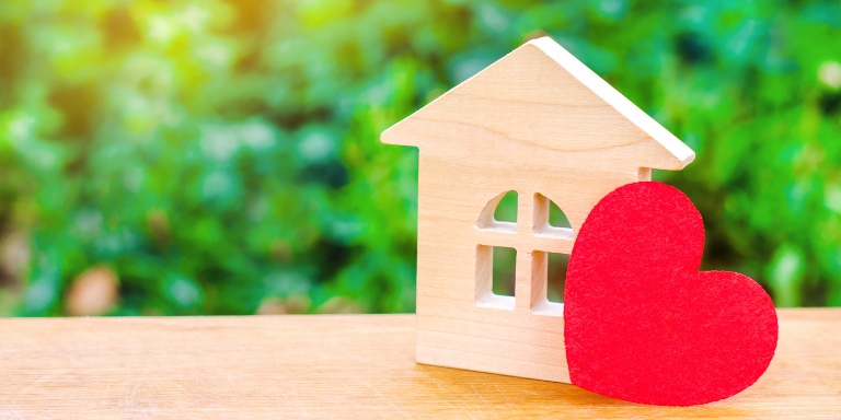 Wooden figurine of a house and a red heart on a table with a blurred background of trees.
