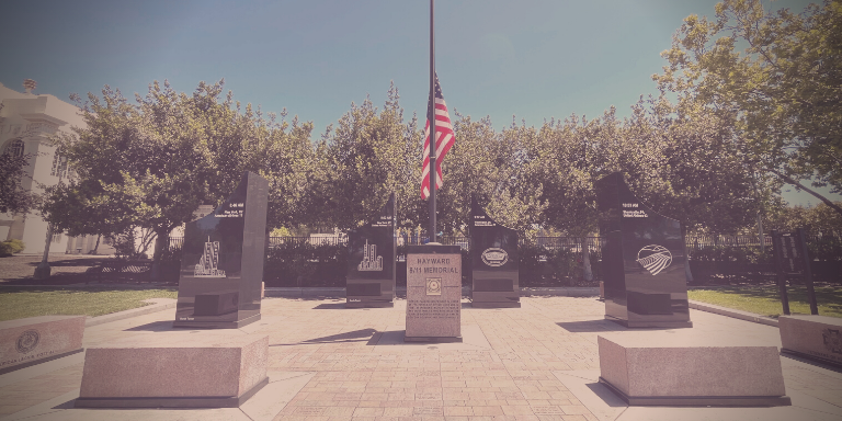 Photo of the Hayward 9/11 Memorial, a paved space with four large black rectangular structures with engravings. In the center, a brown rectangular block with the US flag rising from it.