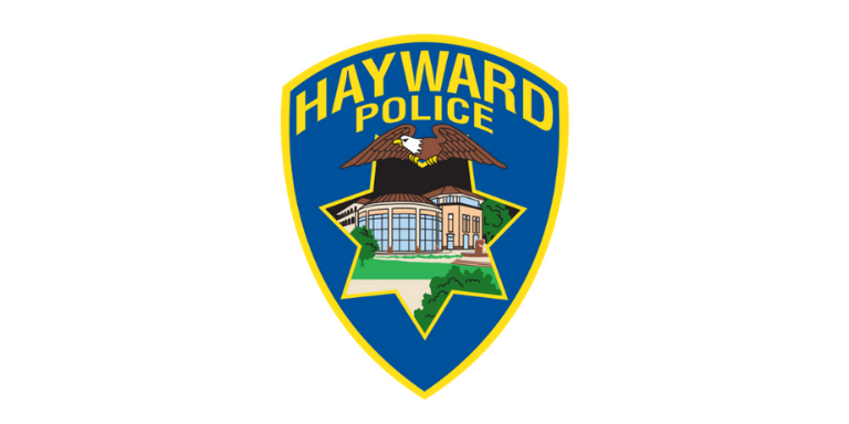 Hayward Police Logo in the shape of a blue badge with Hayward Police written across the top in yellow. An illustration of a Bald Eagle over an illustration of City Hall in the shape of a 7-point star is in the middle.