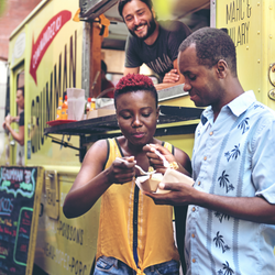Photo of two Black people eating food they ordered from a food truck behind them. A Latino man smiles in the truck behind them.