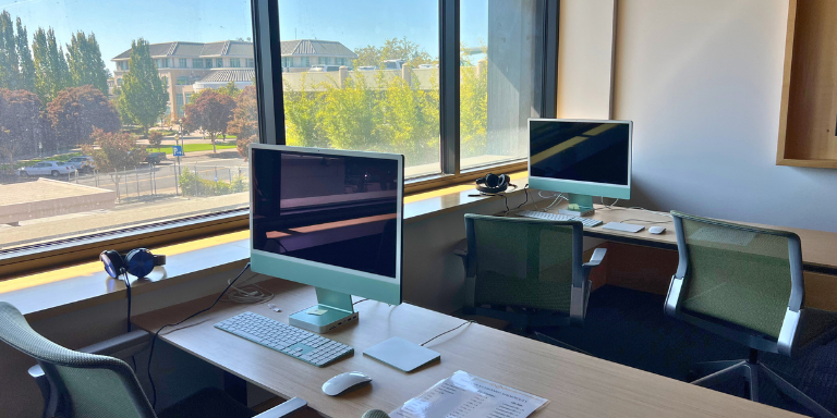 Two iMac computers at desks next to a window.