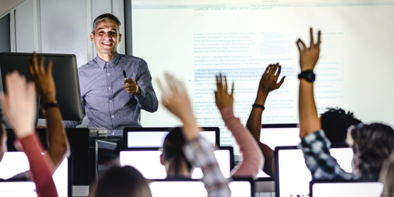 Man leads computer class in front of projector screen, students at computers with hands raised.