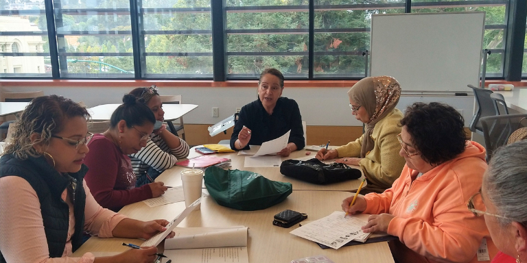 Instructor sits at table with 6 students, students are taking notes.