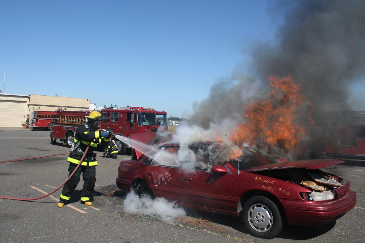 A fire fighter recruit in full turnout gear practices extinguishing a car fire