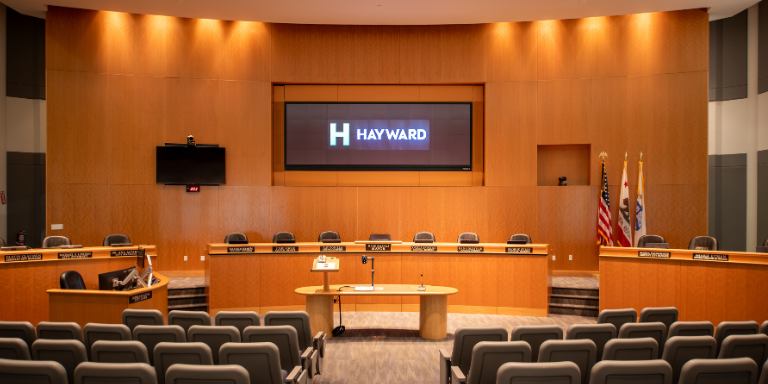 Photo of the City of Hayward's City Council Chamber. An auditorium with wood panelling, a wooden dais, audience chairs, and a large screen at the front.