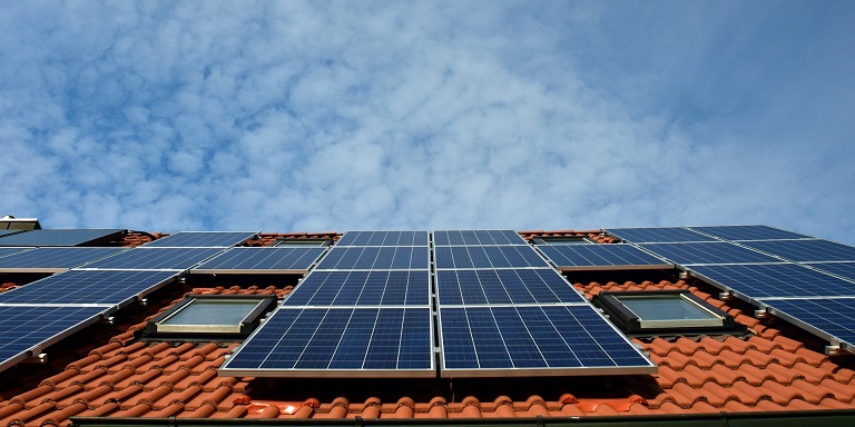 solar panels on a red-orange Spanish tile roof against a blue-partly cloudy sky.