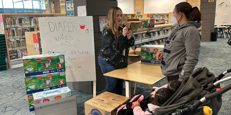 Diaper Kit giveaway at the library. A woman writing on a post-it speaking to a mother of two.