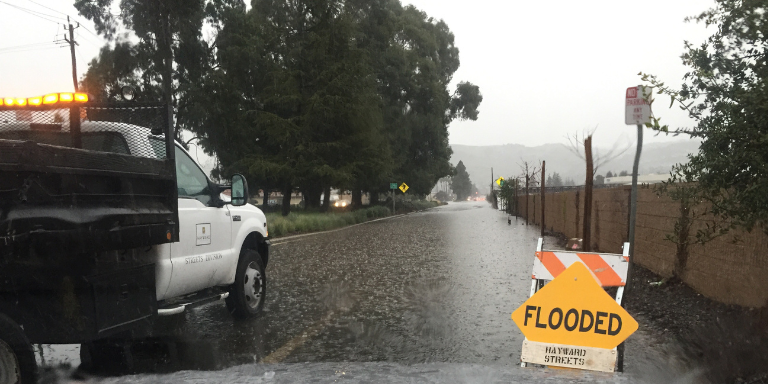 Photo of a City of Hayward truck in the rain next to a sign that reads: Flooded.