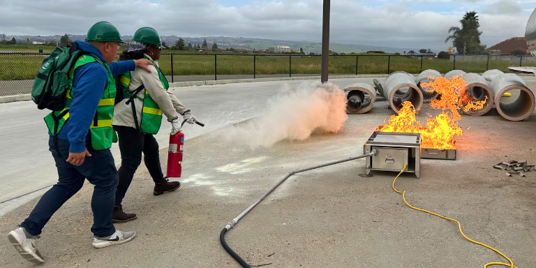 Two people in safety vests and hats holding a fire extinguisher putting out a demo-fire.