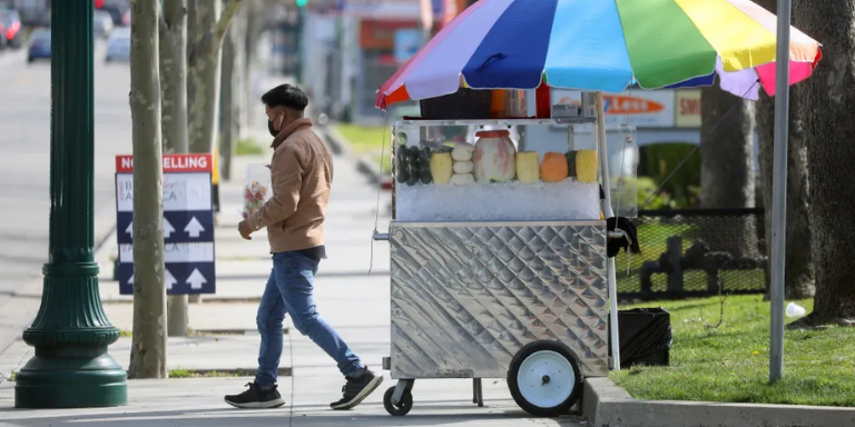 Photo of a man walking away from a fruit cart on a sidewalk.