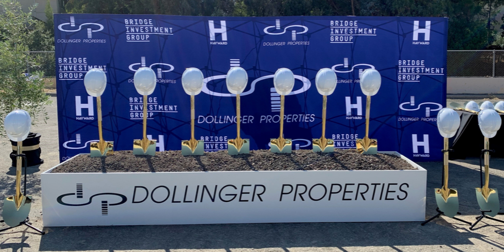 Gold shovels with white hard hats lined up in front of a blue back drop with City of Hayward, Bridge Investment Group and Dollinger Properties logos in white