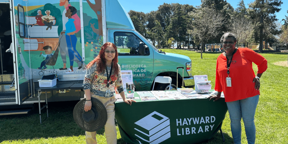 Two members of library staff standing near a library table in front of Curbie, the mobile library branch at an event in a park
