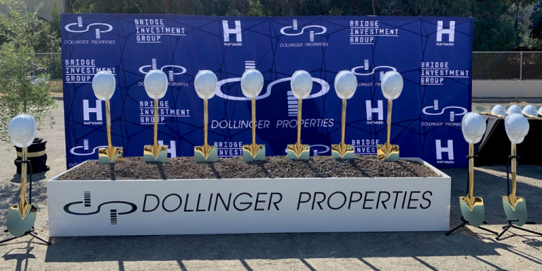 Gold shovels and white hard hats propped up in dirt in front of a blue sign with white Hayward, Dollinger Properties and Bridge Investment Group logos. 