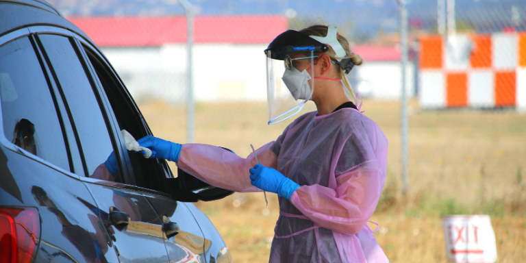 Woman in PPE next to a car administering a COVID-19 test.