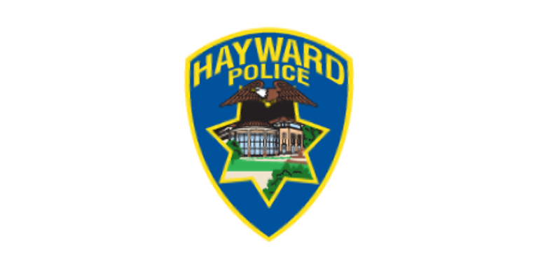 Hayward Police Logo in the shape of a blue badge with Hayward written across the top in yellow. An illustration of a Bald Eagle over an illustration of City Hall in the shape of a 7-point star is in the middle.