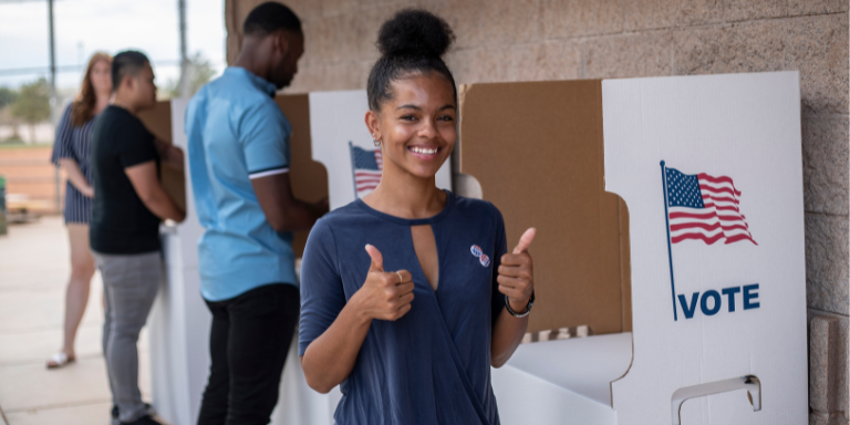 A Black woman with two thumbs up in front of a voting booth. In the background, other people at a voting booth.