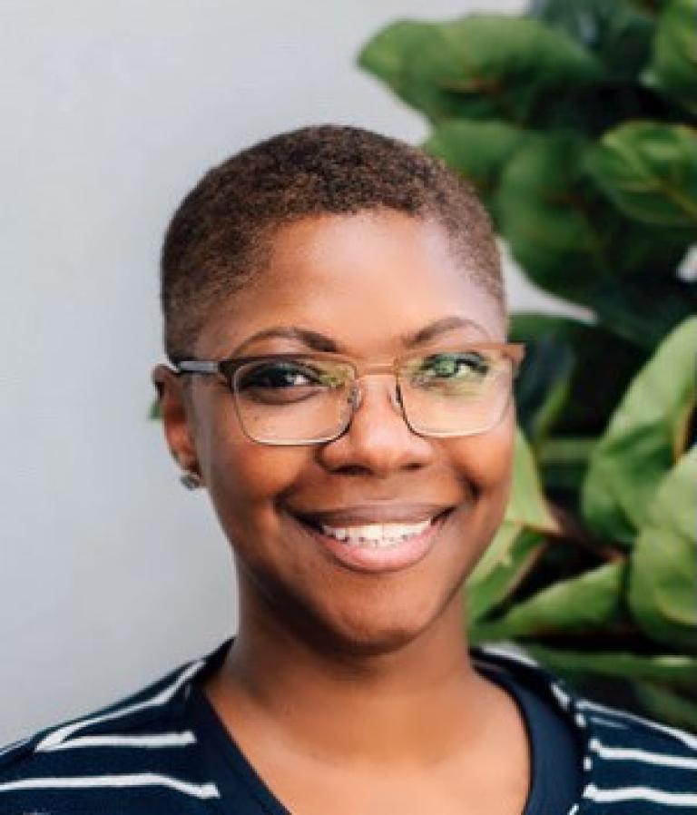 Professional headshot of a smiling Black woman with short hair and glasses with a green plant in the background.