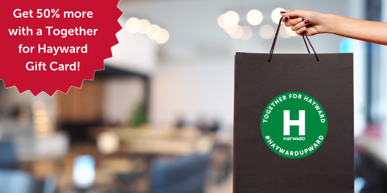 Photo of a person holding a black shopping bag with the Together for Hayward logo on it. In the top left corner on a red circle with white text that reads Get 50% more with a Together for Hayward Gift Card!
